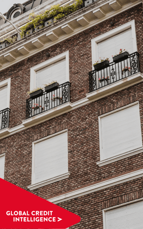 Moderno edificio comercial de ladrillo con contraventanas cerradas y balcones estructurados, con una pancarta roja superpuesta en la que se lee "Global Credit Intelligence", que representa información sobre los mercados inmobiliario y crediticio.