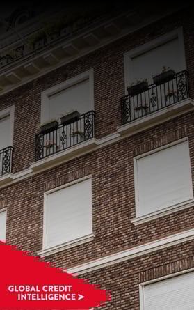 Moderno edificio comercial de ladrillo con contraventanas cerradas y balcones estructurados, con una pancarta roja superpuesta en la que se lee "Global Credit Intelligence", que representa información sobre los mercados inmobiliario y crediticio.