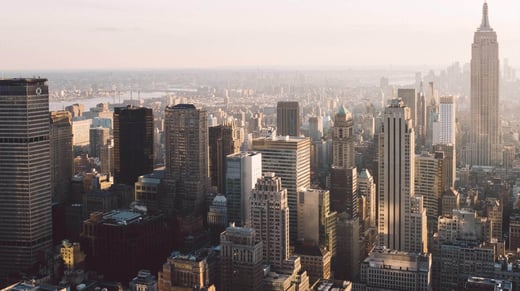 Vista aérea del horizonte de Nueva York al atardecer, con numerosos rascacielos y edificios altos. El Empire State Building se ve a la derecha, bañado en una cálida luz dorada, con un brumoso paisaje urbano que se extiende en la distancia.