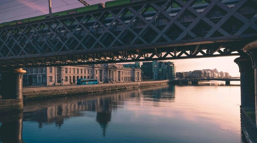 Una vista panorámica del río Liffey en Dublín, Irlanda, al atardecer, con el histórico puente Loopline y un tren pasando por encima. Abajo, las aguas tranquilas reflejan las estructuras circundantes. El edificio neoclásico Custom House bordea el río, con modernos edificios de cristal al fondo. En la calle se ve un autobús, que contribuye al ambiente urbano.