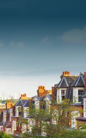 Vista panorámica de casas victorianas de ladrillo rojo en Highgate, Londres, con sus características chimeneas y ventanas abuhardilladas. La exuberante vegetación del primer plano contrasta con el moderno horizonte de Canary Wharf, visible en la brumosa distancia.