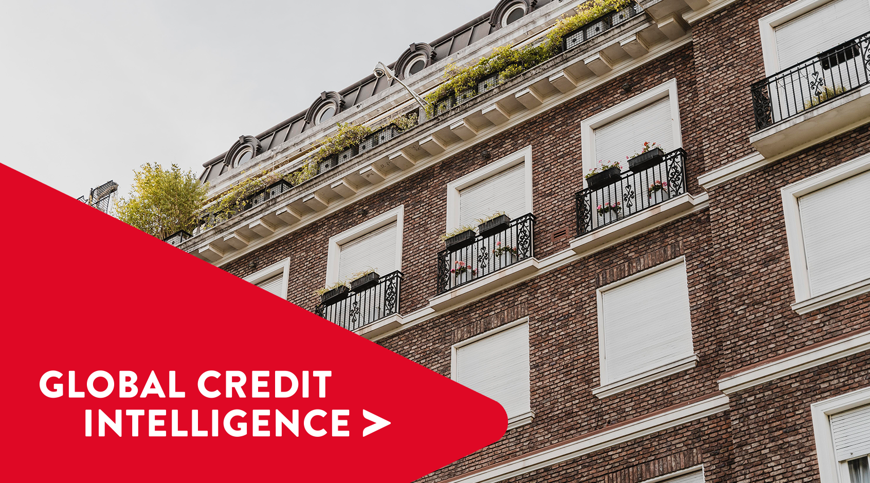 Moderno edificio comercial de ladrillo con contraventanas cerradas y balcones estructurados, con una pancarta roja superpuesta en la que se lee "Global Credit Intelligence", que representa información sobre los mercados inmobiliario y crediticio.