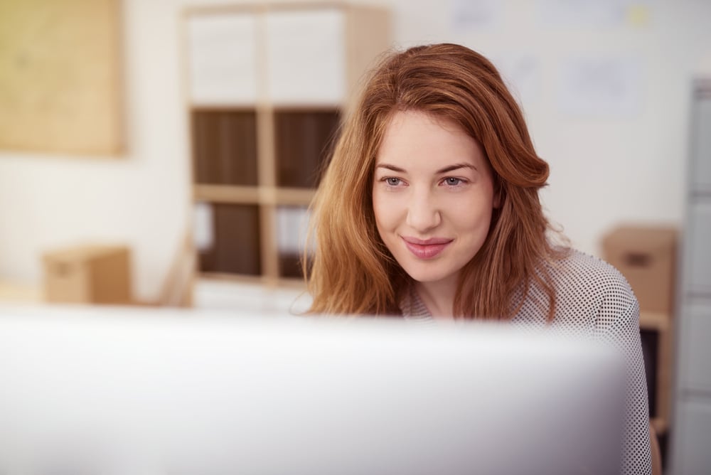Mujer joven y atractiva trabajando en un ordenador de sobremesa sonriendo mientras se inclina hacia delante leyendo un texto en la pantalla, vista sobre el monitor